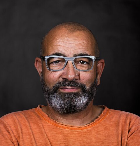 Roland Rudolf | © wob AG Dieses Bild ist ein Porträt von Roland Rudolf. Er hat einen Bart und eine Brille. Er trägt ein orangenes Sweatshirt. Der Hintergrund ist dunkel. | © wob AG