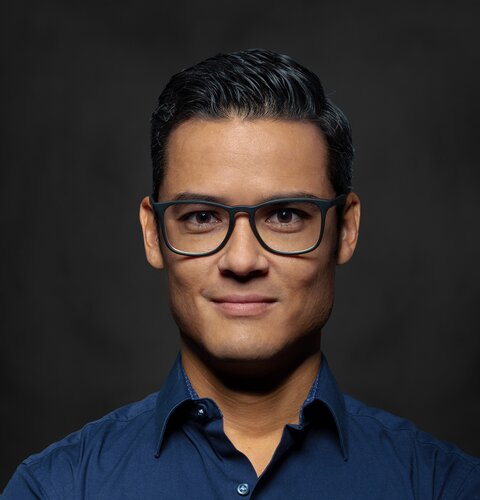 Dennis Güth | © wob AG Dieses Bild ist ein Porträt von Dennis Güth. Er trägt eine Brille und ein dunkel blaues Hemd. Seine Arme sind verschränkt. Der Hintergrund ist dunkel. | © wob AG