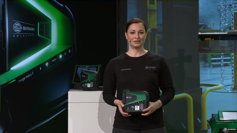 #ZUHAUSEMESSE Werkstattshow mit Lina van de Mars Dieses Bild zeigt einen Screenshot aus dem digitalen Live-Event. Man sieht die Moderatorin Lina van de Mars bei der Moderation des digitalen Live -Events #Zuhausmesse. Sie trägt ein schwarzes Longsleeve-Shirt und die Haare im Nacken zusammengebunden und blickt in die Kamera. Sie hält einen mega macs X in ihren Händen. Hinter ihr befindet sich ein Moderatorenpult und ein großer Screen mit dem Kampagnen-Hauptmotiv, einer hochwertigen Produktabbildung auf schwarzem Hintergrund.