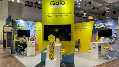 Messestand GoTo Das Foto zeigt einen Messestand von GoTo. Das ist daran zu erkennen, dass sich das Logo auf allen Messewänden widerspiegelt. Eine Dame sitzt an einem Tisch. Zu sehen sind noch weitere Tische, die Gesprächssituationen am Messestand erlauben sollen.