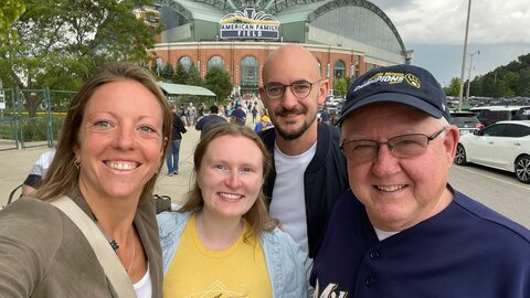 BBN-Austausch Auf einem Foto sind 4 Personen zu sehen, die fröhlich in die Kamera lächeln. Darunter sind zwei junge Frauen, ein junger Mann und ein etwas älterer Herr mit Baseballkappe. Hinter den vier Personen ist ein Baseballstadion zu erkennen.